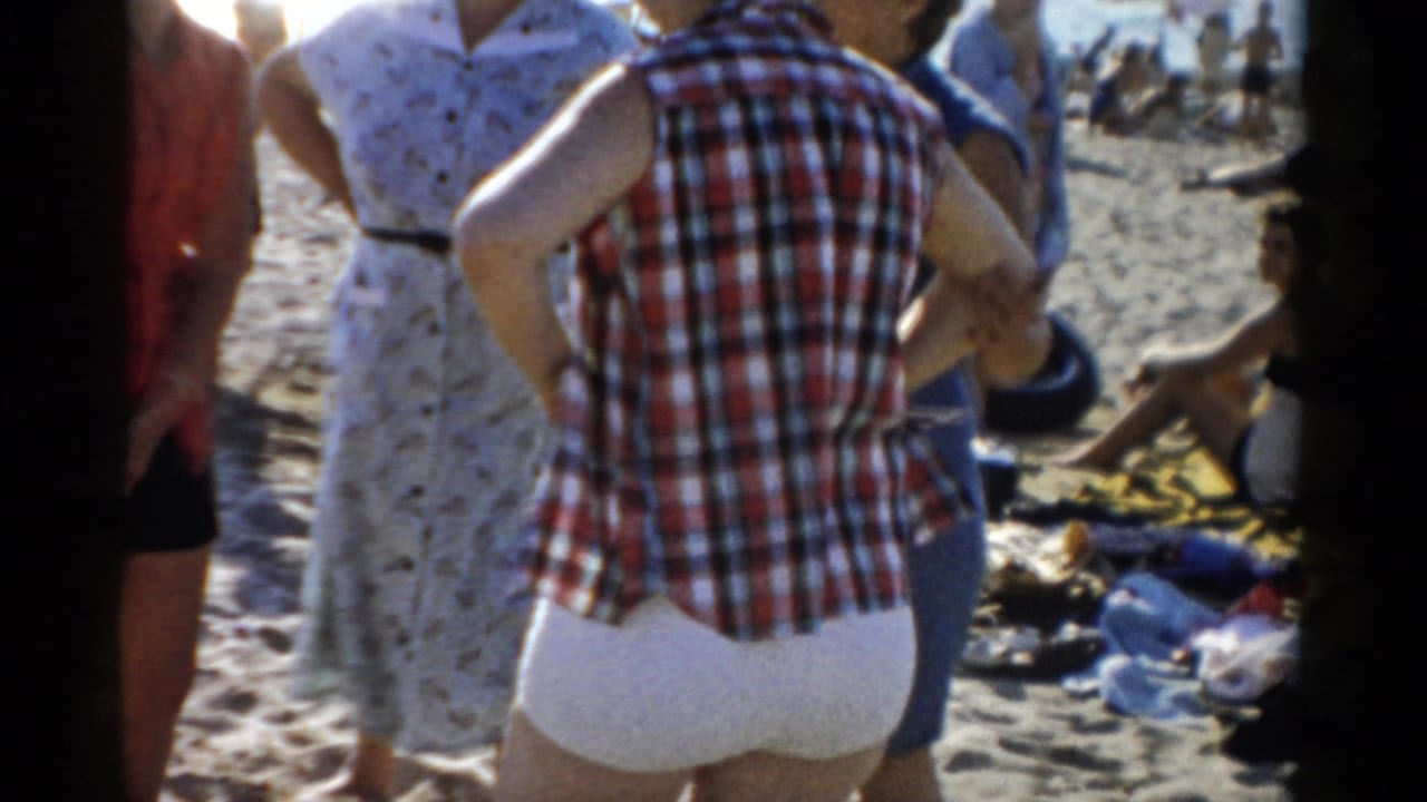 Person in red-and-white plaid shirt and white shorts stands on a crowded beach seen from behind, with other beachgoers and scattered belongings on sand.