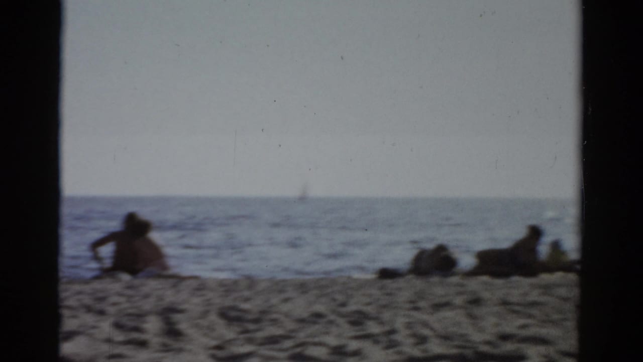 Wide shot of people lounging near the waterline with calm water and a distant possible sailboat; Catskill, New York, circa 1957 8mm home movie.
