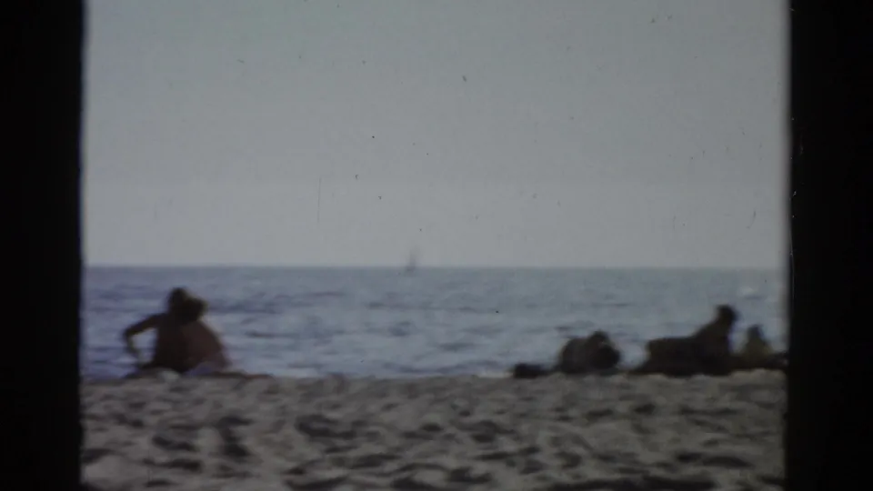 Wide shot of people lounging near the waterline with calm water and a distant possible sailboat; Catskill, New York, circa 1957 8mm home movie.