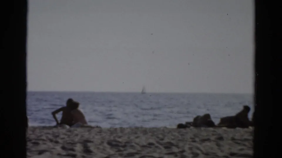 Wide beach shot with seated figures silhouetted against calm water and a pale sky; Catskill, New York, circa 1957 8mm home movie.