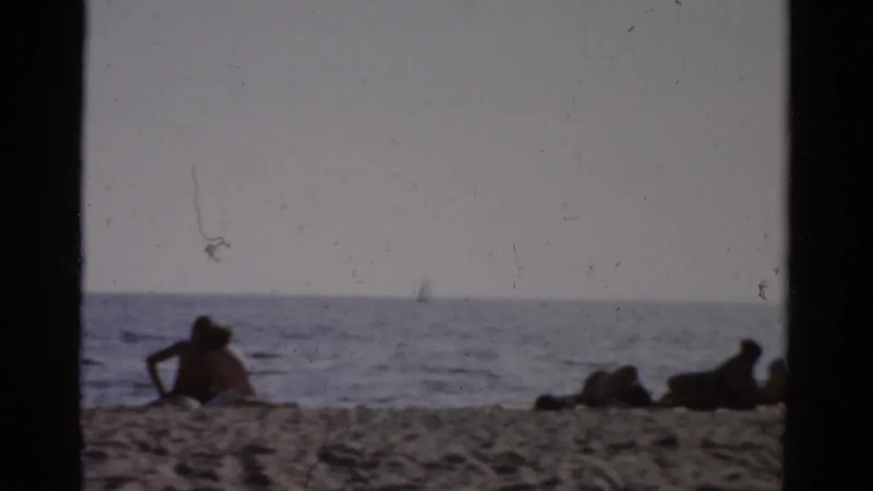 Beachgoers resting on sand near calm water with a distant possible sailboat; Catskill, New York, circa 1957 8mm home movie.