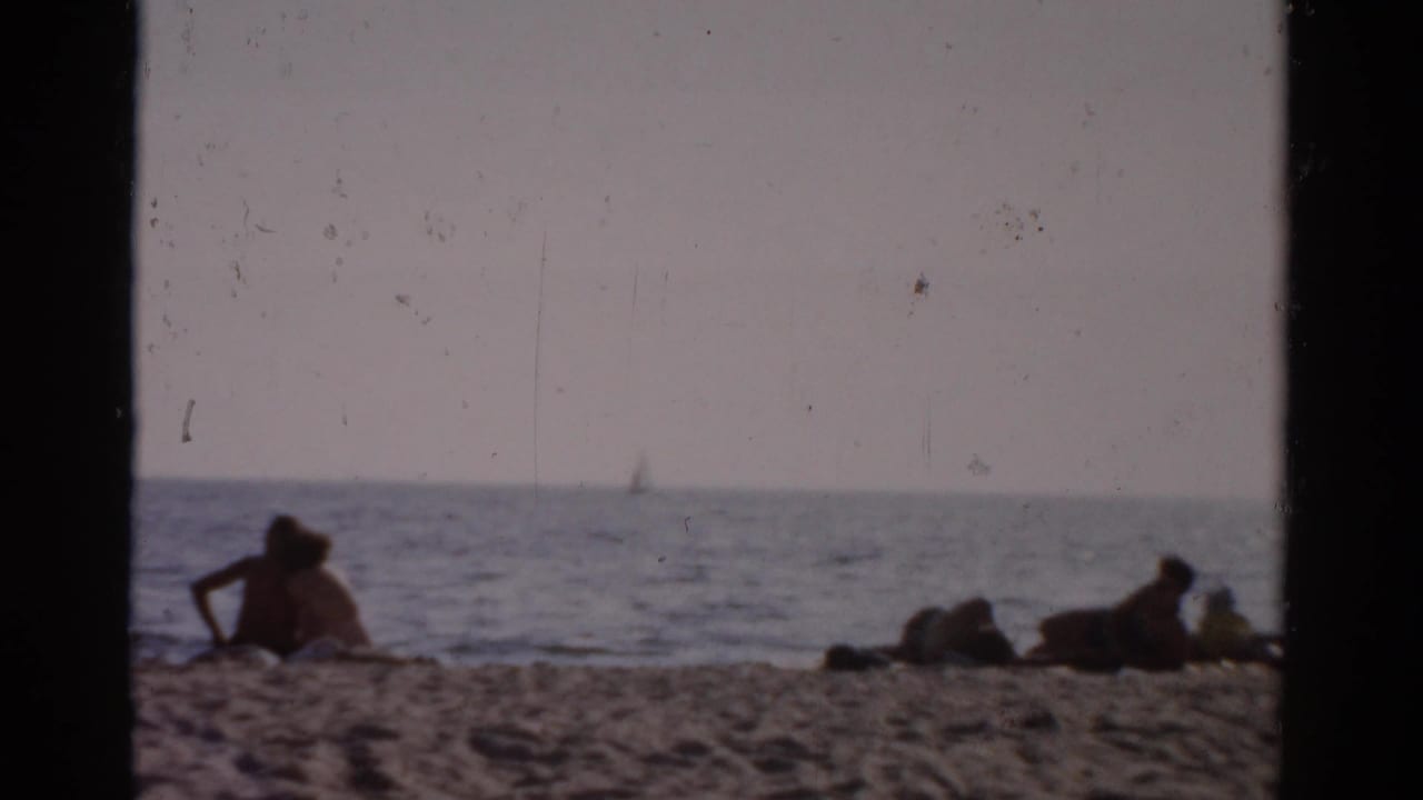 Sunbathers on a sandy beach, likely upstate New York circa 1957, with a distant sailboat on calm water under a hazy sky.