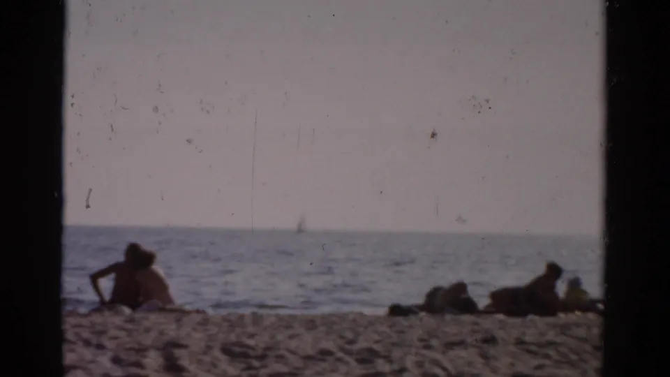 Sunbathers on a sandy beach, likely upstate New York circa 1957, with a distant sailboat on calm water under a hazy sky.
