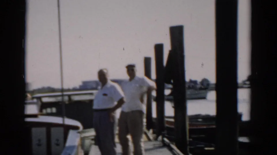 Two men in white shirts on a wooden dock, likely near Catskill, New York circa 1957, with dock pilings and a moored boat nearby.