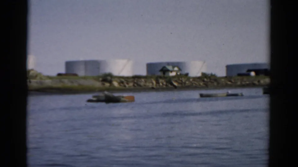 Industrial waterfront with large white cylindrical storage tanks across calm harbor water, possibly near Catskill, New York, circa 1957.
