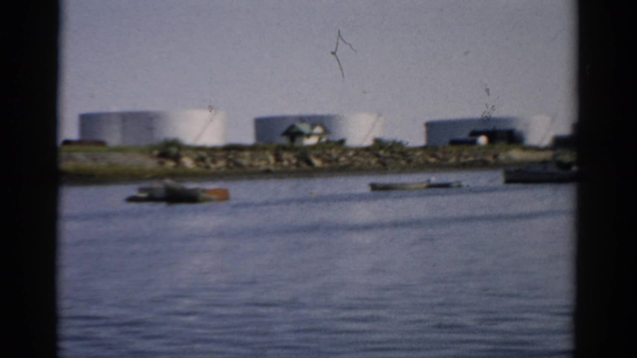 Catskill, New York waterway, c.1957 — small boats moored near a low rocky shoreline, white storage tanks and industrial buildings in background, 8mm home movie.