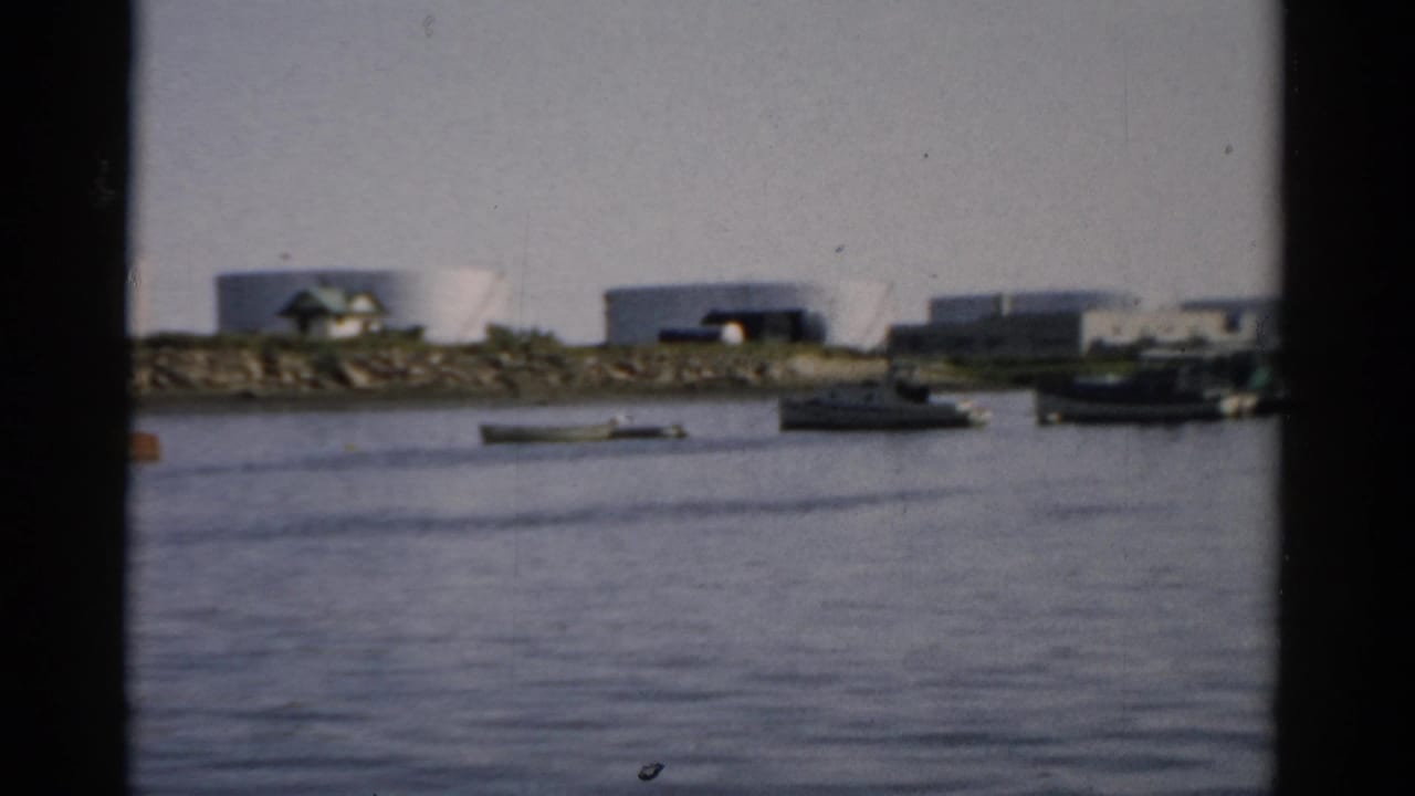 Same waterway scene with moored boats and white storage tanks; a red-orange element visible among the boats, slight camera shift, 8mm film.
