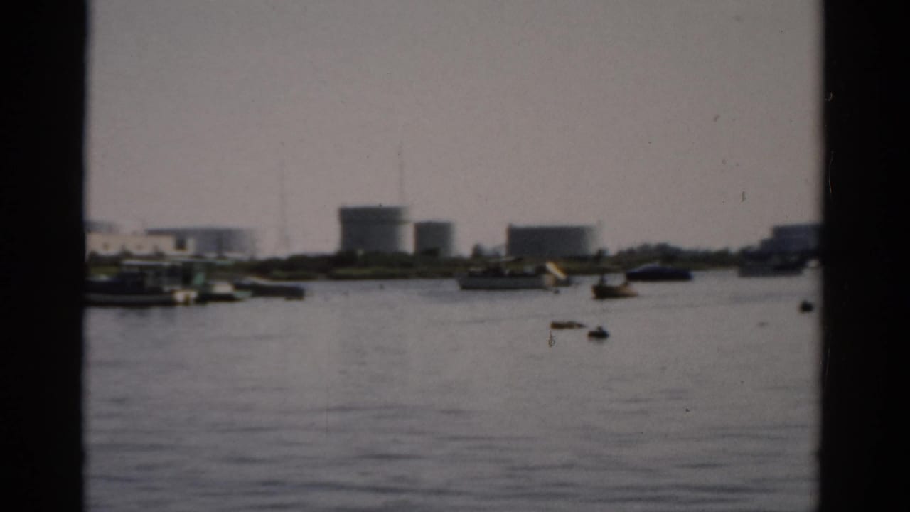 Calm harbor water with scattered small boats and cylindrical industrial tanks on the distant shore, shot through a window frame on 8mm film.