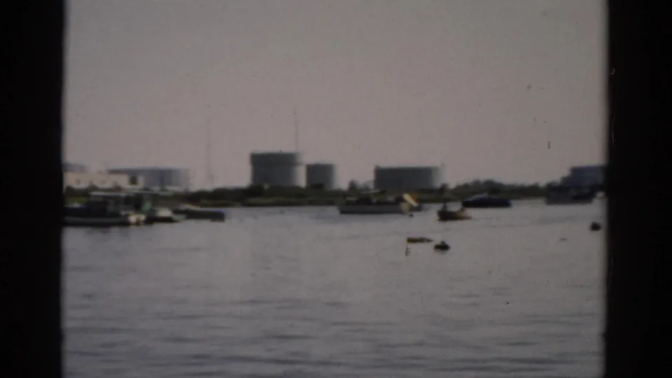 1957 8mm view across a calm harbor in Catskill, New York, showing small boats on flat water and cylindrical storage tanks on the far industrial shoreline.