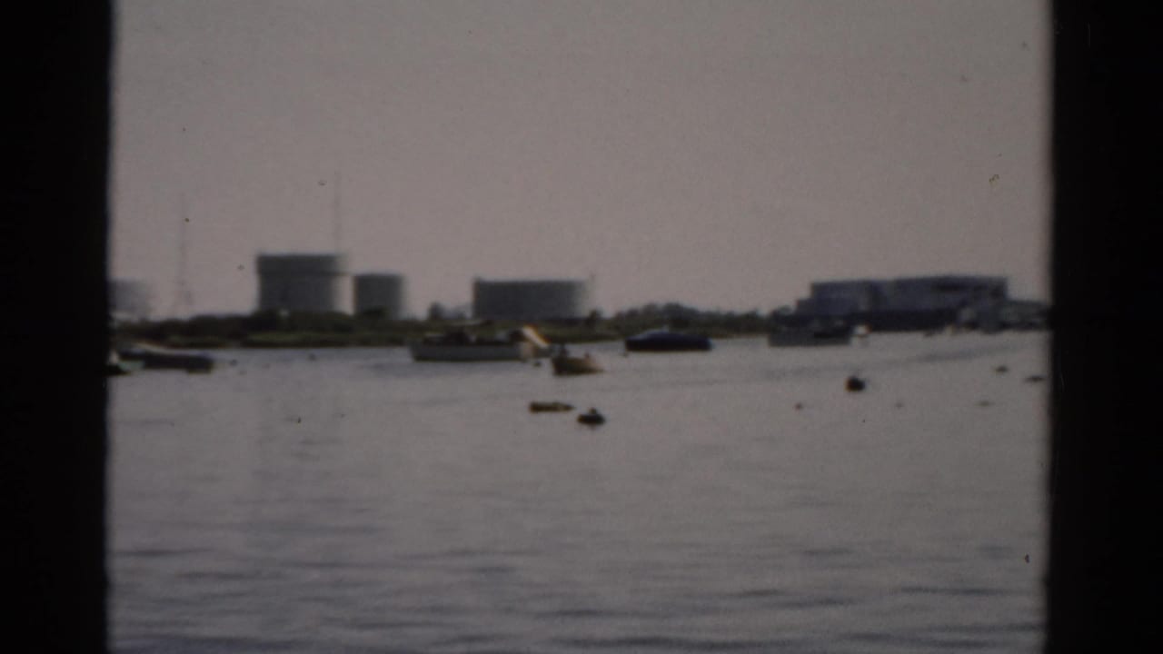 8mm frame circa 1957 showing Catskill, NY harbor with industrial storage tanks and moored small boats under a hazy overcast sky.