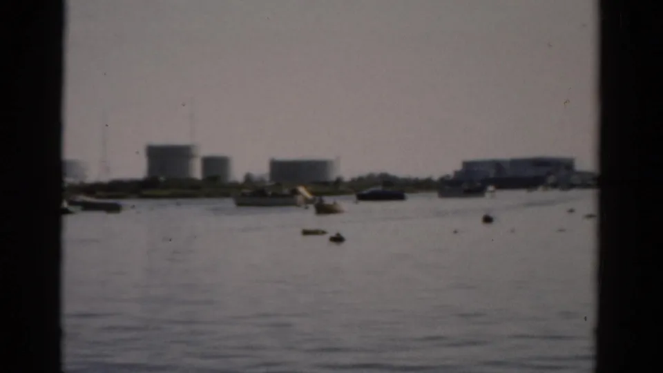 8mm frame circa 1957 showing Catskill, NY harbor with industrial storage tanks and moored small boats under a hazy overcast sky.