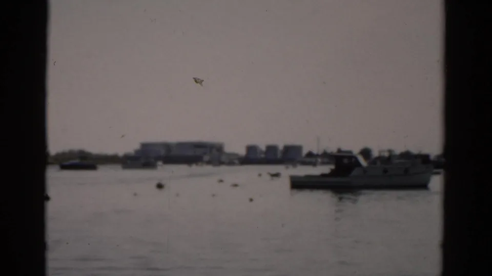 Final 8mm frame circa 1957 from Catskill, NY showing a motorboat and smaller vessels on calm harbor water with white buildings on the distant shore.