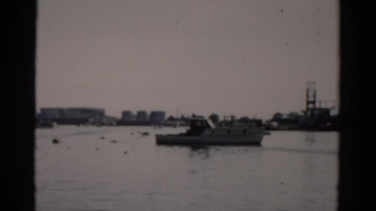 Harbor view with moored boats on calm water, industrial buildings and a tall structure visible on the far shore.