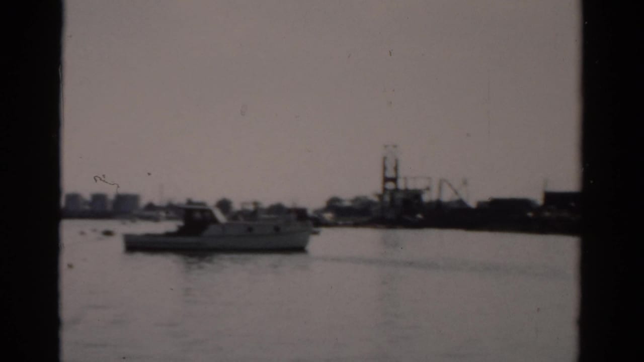 1950s harbor, possibly Catskill, New York — calm water, white motorboat at moorings, low industrial skyline, and a crane structure in the background.