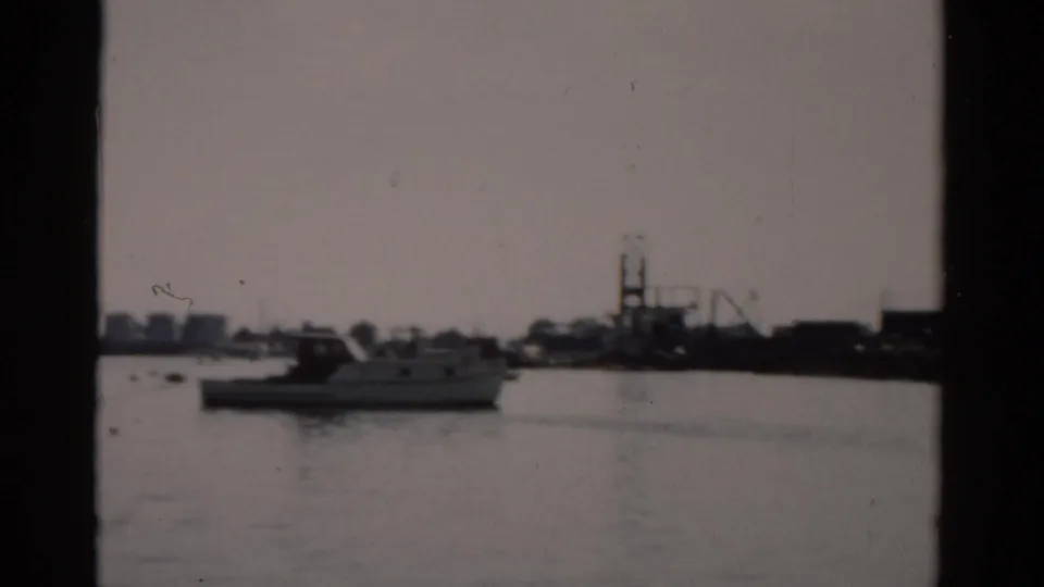 1950s harbor, possibly Catskill, New York — calm water, white motorboat at moorings, low industrial skyline, and a crane structure in the background.