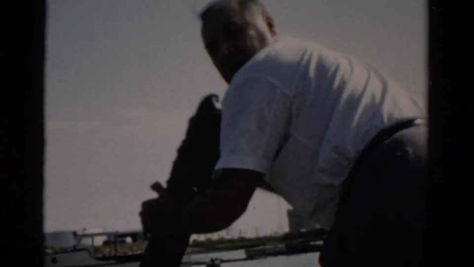 Man in 1950s-style white shirt and dark trousers leaning over boat rail or deck equipment; harbor with moored vessels visible behind him, circa 1957.