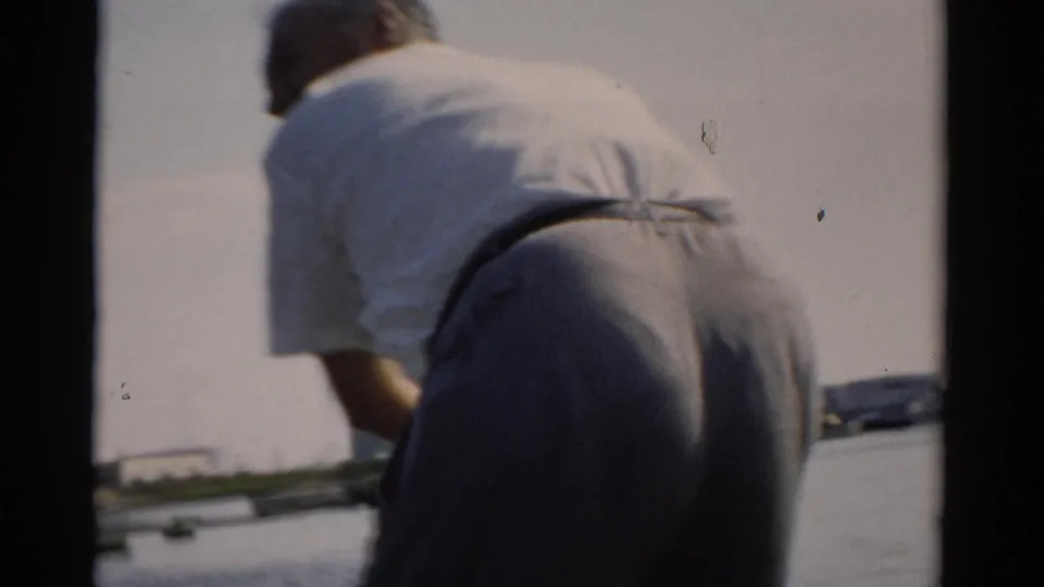 Close view of man in 1950s casual attire bent over boat deck, seen from behind; white deck surface and pale horizon visible, circa 1957.