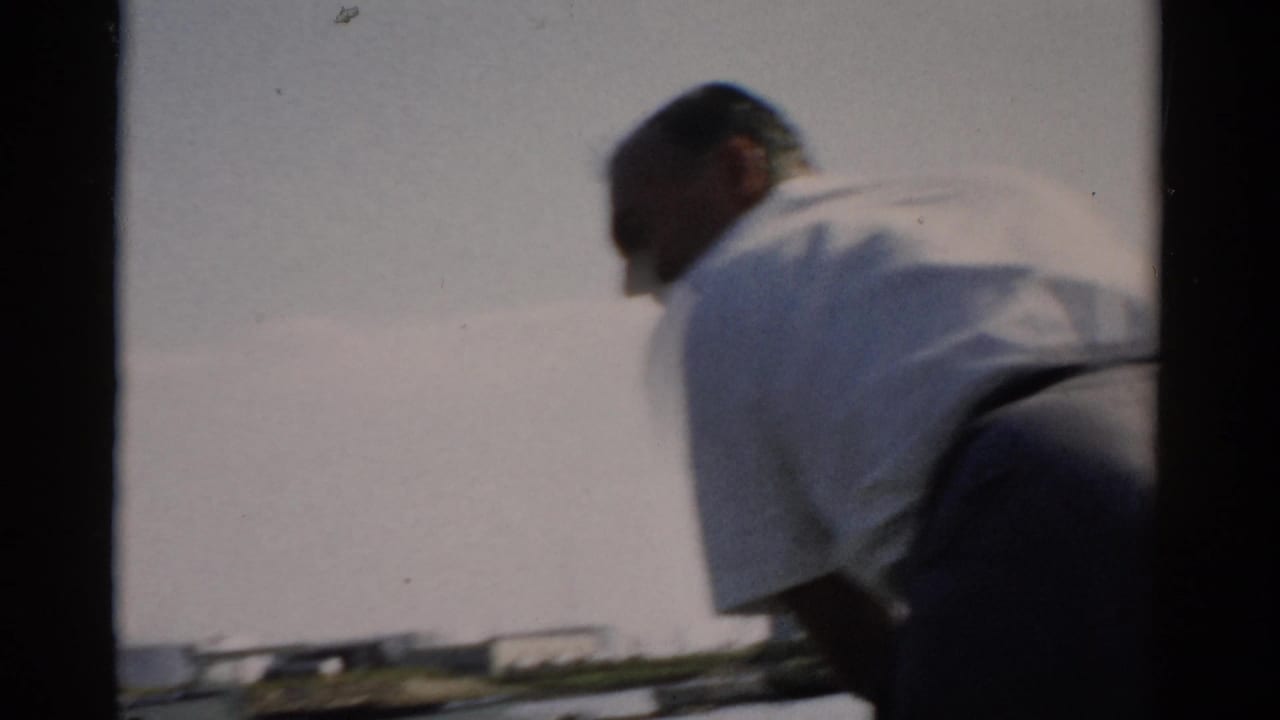 Back view of a heavyset man in a white shirt and blue cap seated on a small boat, with calm water and a distant shoreline behind him.
