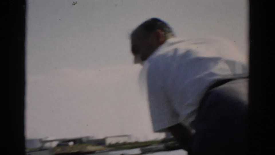 Heavyset man seen from behind in a white short-sleeve shirt and blue cap on a small boat on a lake, likely Catskill, New York, circa 1957, shot on 8mm film.