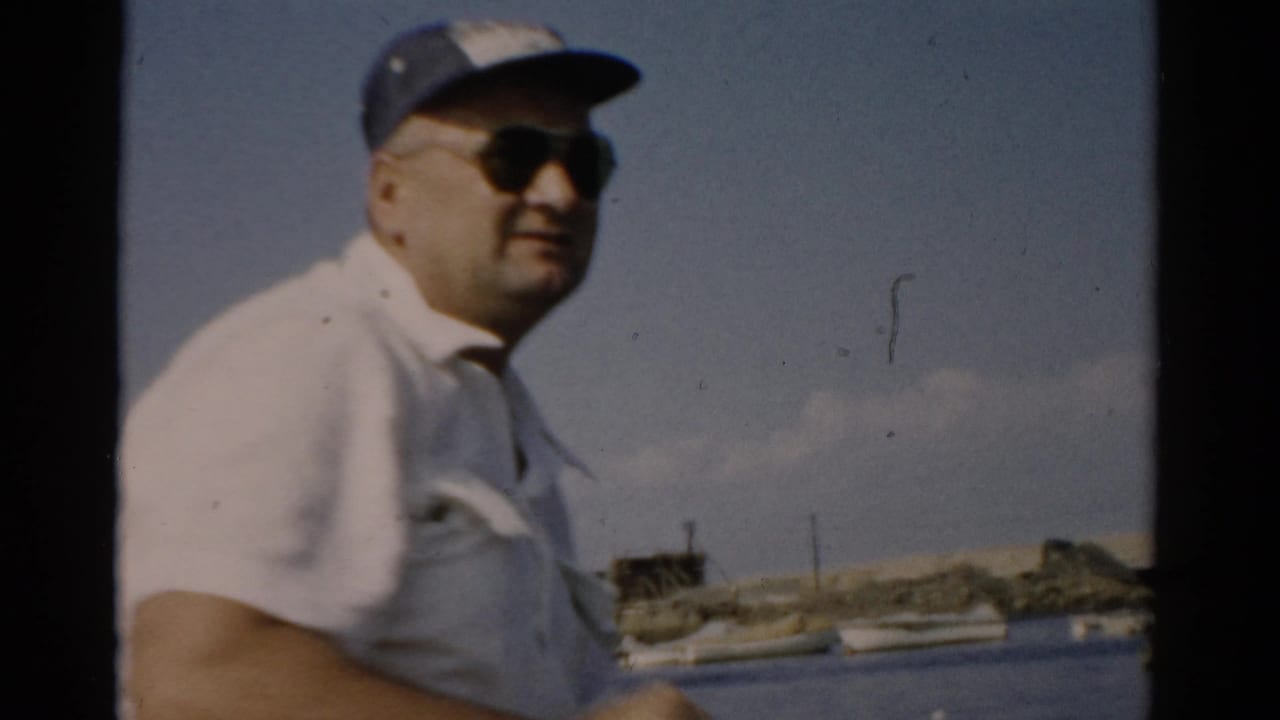 Close view of a heavyset man wearing dark sunglasses and a blue cap on a boat, with calm lake water and a distant waterfront behind him.