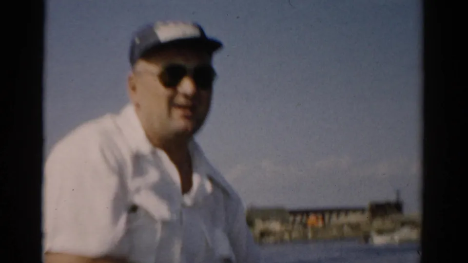 Man wearing dark sunglasses, blue cap, and white shirt turns toward the camera while in a small rowboat on a calm lake, likely Catskill, New York, circa 1957.