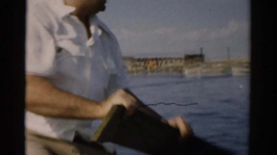 Man gripping what appears to be a makeshift wooden plank used as an oar while in a small boat on a calm lake, with a distant waterfront in the background, circa 1957 Catskill, New York.