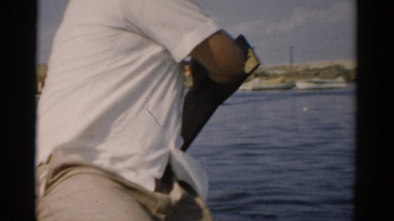 Close view from behind of an older man in a white shirt working a hand pump from a small boat, calm water and a low shoreline behind him; 8mm home movie, Catskill NY area, c. 1957.