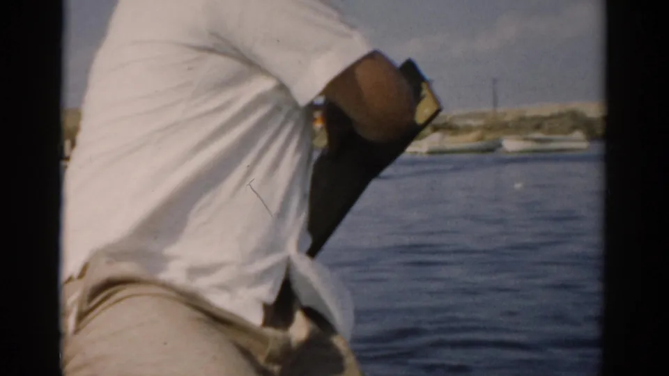 Close view from behind of an older man in a white shirt working a hand pump from a small boat, calm water and a low shoreline behind him; 8mm home movie, Catskill NY area, c. 1957.