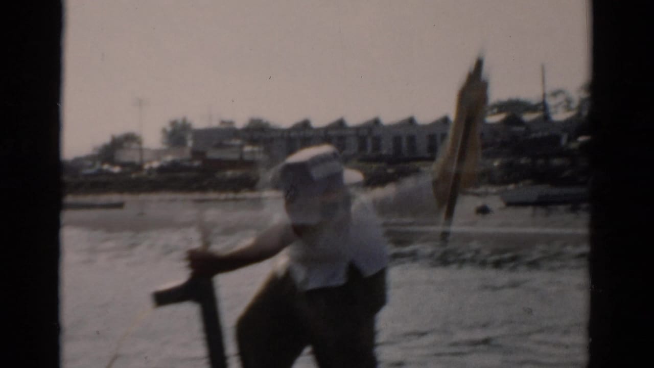 Older man in white short-sleeved shirt and light cap works a hand pump aboard a small boat; structures and moored vessels visible on the far shore; 8mm home movie, c. 1957.