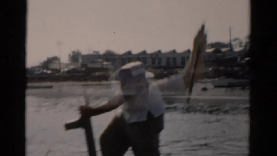 Older man in white short-sleeved shirt and light cap works a hand pump aboard a small boat; structures and moored vessels visible on the far shore; 8mm home movie, c. 1957.