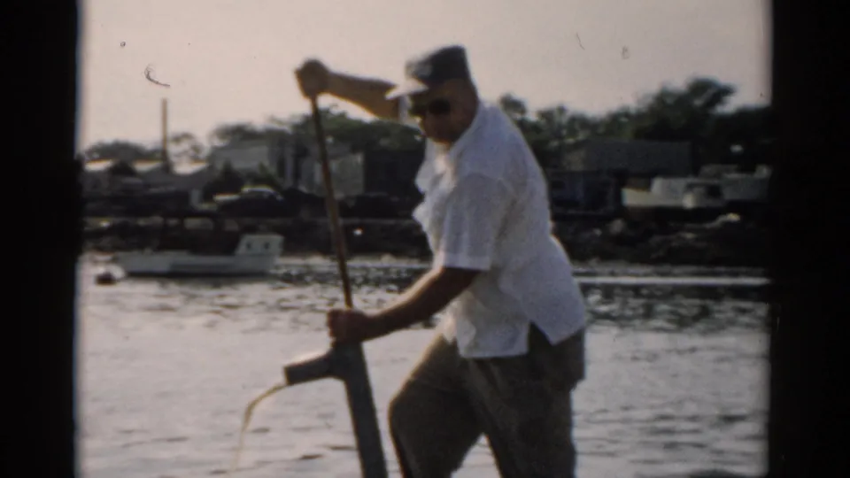 Older man in white short-sleeved shirt, dark sunglasses, and dark cap vigorously works a manual bilge pump; water flows from the outlet; trees and structures on far shore; 8mm home movie, c. 1957.