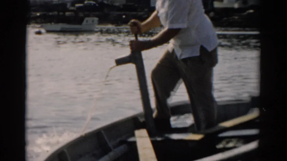 Side view of an older man in a white shirt operating a manual hand pump from a wooden rowboat; boat interior and gunwale visible in foreground; waterfront structures on far shore; 8mm home movie, c. 1957.