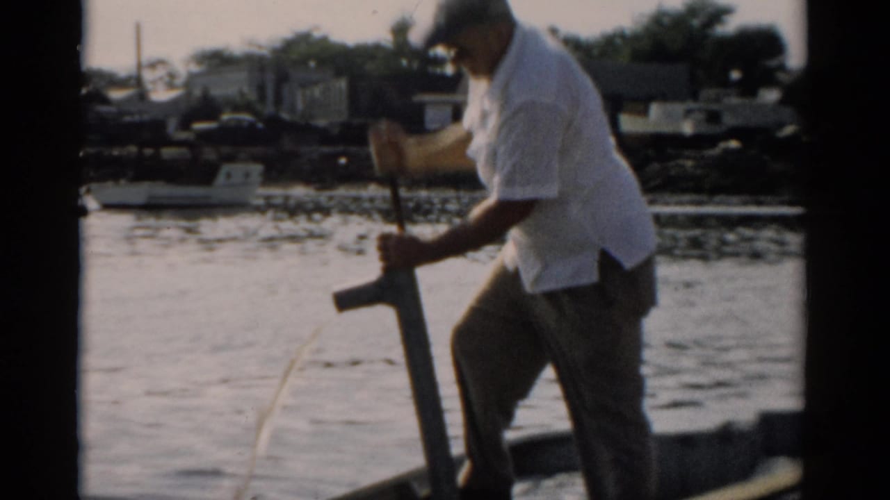 Man in white short-sleeved shirt and dark trousers works a manual bilge pump on a small wooden boat on calm water, Catskill, New York, c. 1957.