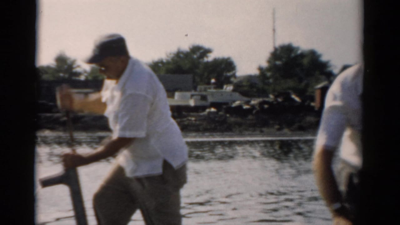 Two men on a small wooden boat on calm water near Catskill, New York, c. 1957; one operates a manual bilge pump, the other stands at right.