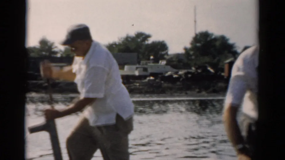 Two men on a small wooden boat on calm water near Catskill, New York, c. 1957; one operates a manual bilge pump, the other stands at right.