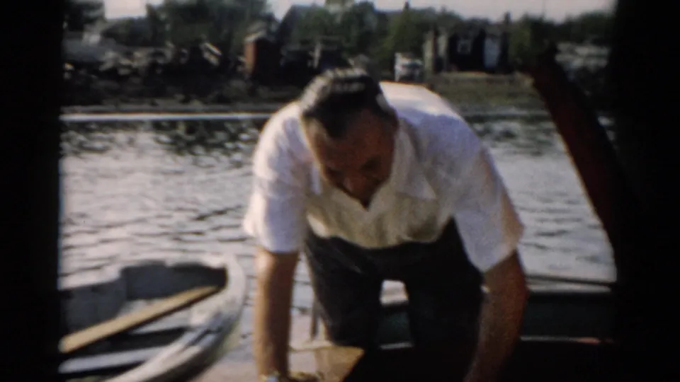 Man in white short-sleeved shirt and dark trousers bends to inspect the interior of a small wooden boat on calm water, likely Catskill, New York, c. 1957.