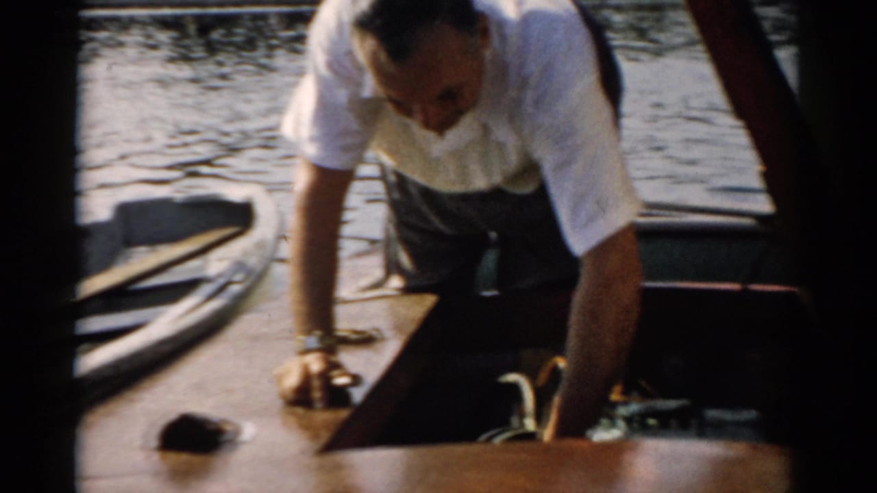 Middle-aged man bends over the varnished deck of a small wooden motorboat near an open engine hatch; water and distant shoreline visible behind, consistent with a 1957 Catskill, New York setting.