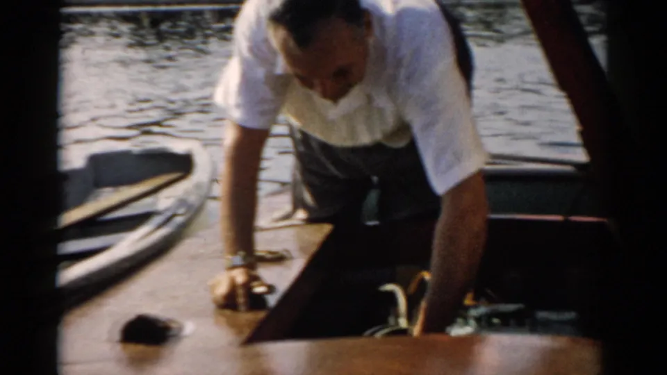 Middle-aged man bends over the varnished deck of a small wooden motorboat near an open engine hatch; water and distant shoreline visible behind, consistent with a 1957 Catskill, New York setting.