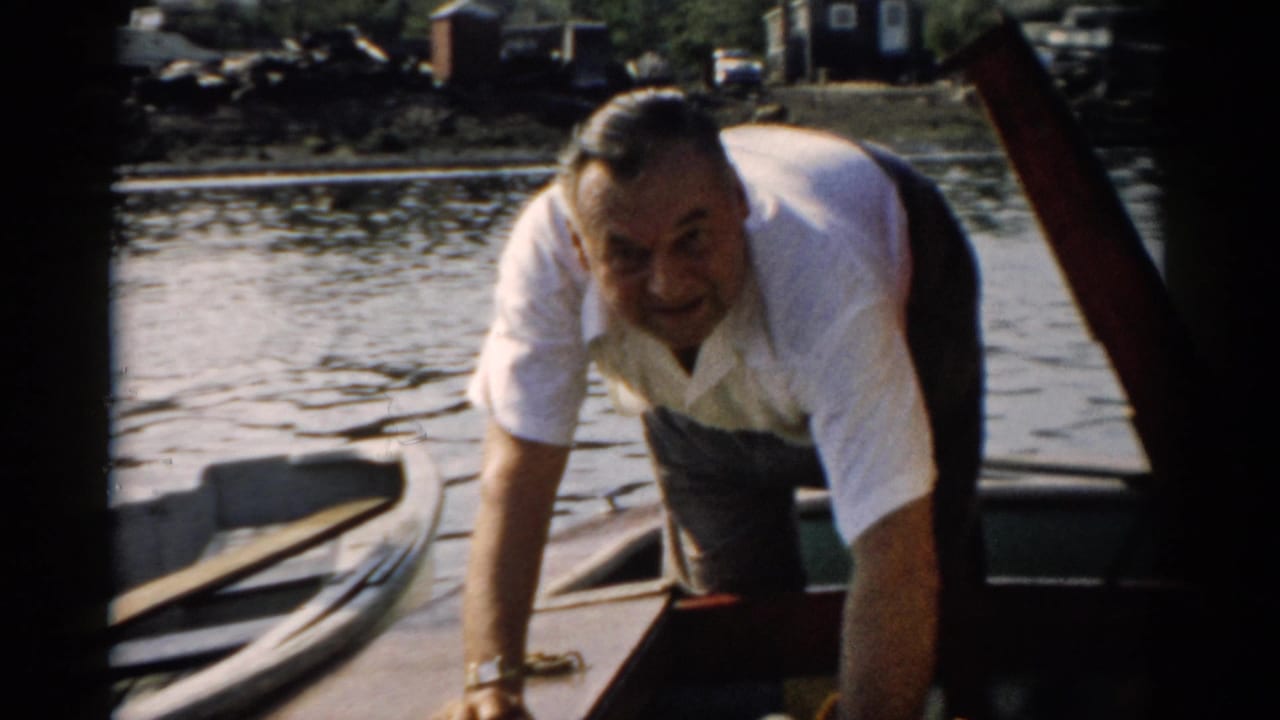 Middle-aged man looks directly at the camera while leaning over a small wooden boat; a rocky bank and distant structures are visible behind, consistent with a 1950s upstate New York waterway.