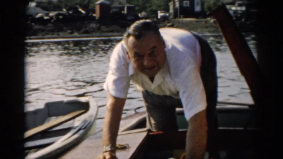 Middle-aged man looks directly at the camera while leaning over a small wooden boat; a rocky bank and distant structures are visible behind, consistent with a 1950s upstate New York waterway.