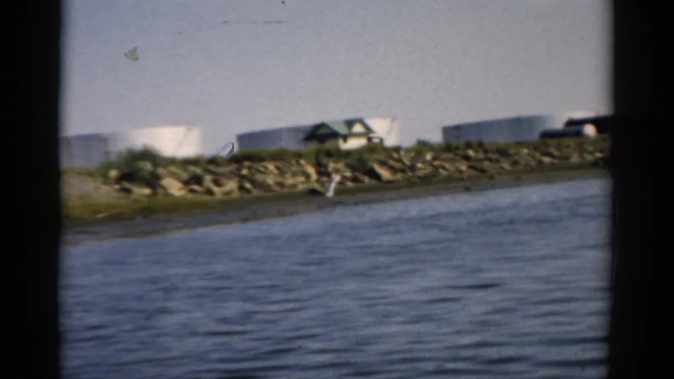 View across calm water toward a rocky, debris-strewn bank; large white cylindrical storage tanks and a small building with green roof are visible on the far shore, possibly Catskill, New York, circa 1957.