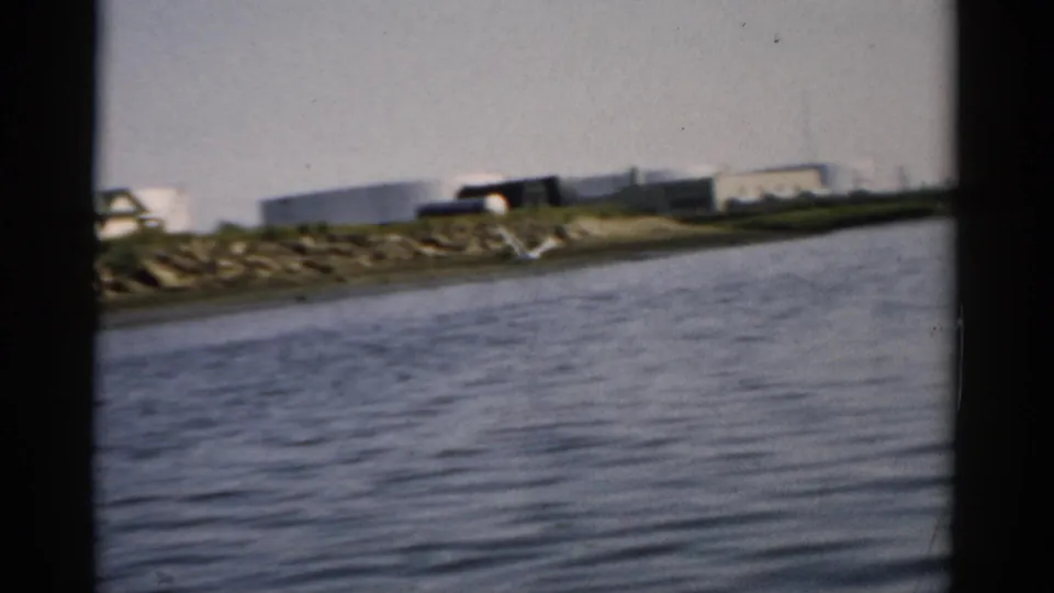 Continued view across calm water toward a rocky shoreline; cylindrical storage tanks and a small building remain visible on the far bank, consistent with footage labeled Catskill, New York, 1957.