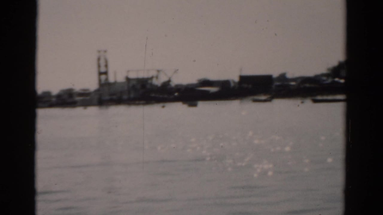 View across flat river water toward a shoreline with crane silhouettes and industrial structures, 8mm film circa 1957.