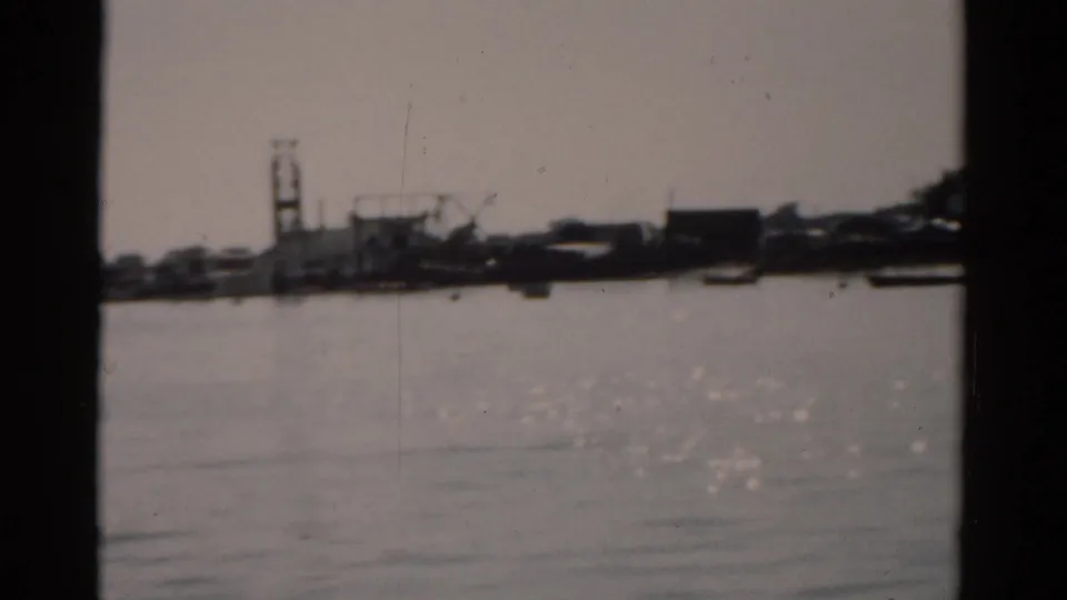View across flat river water toward a shoreline with crane silhouettes and industrial structures, 8mm film circa 1957.