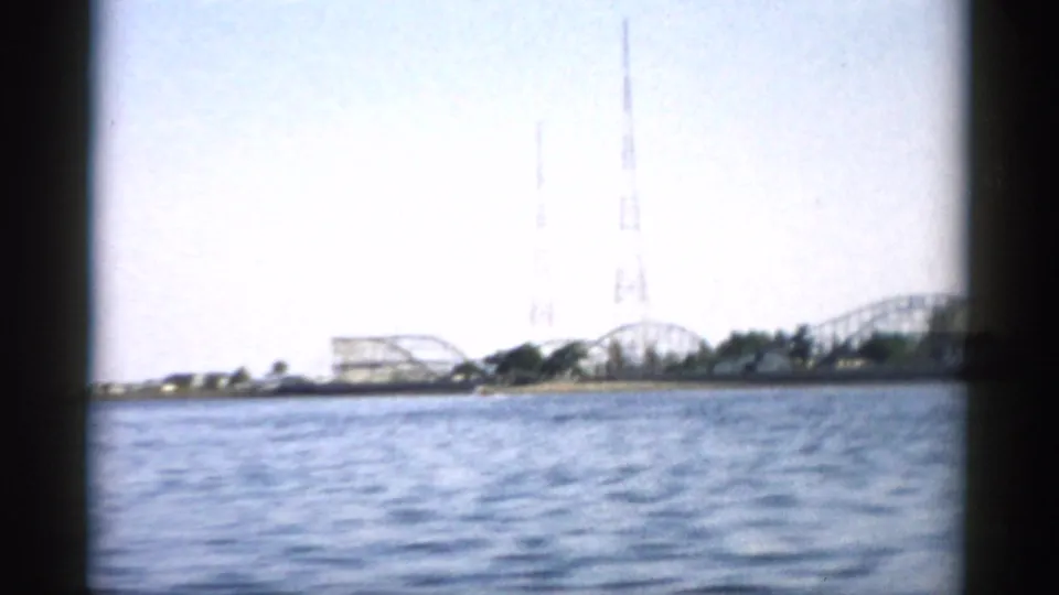 View from water toward a shoreline featuring a prominent tall lattice tower and possible amusement-park structures, 8mm footage circa 1957.