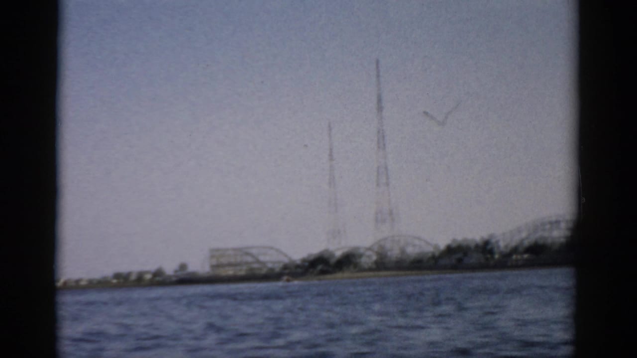8mm home-movie frame c.1957, Catskill-area lake, showing roller coaster and lattice towers on the far shore with a bird crossing the overcast sky.