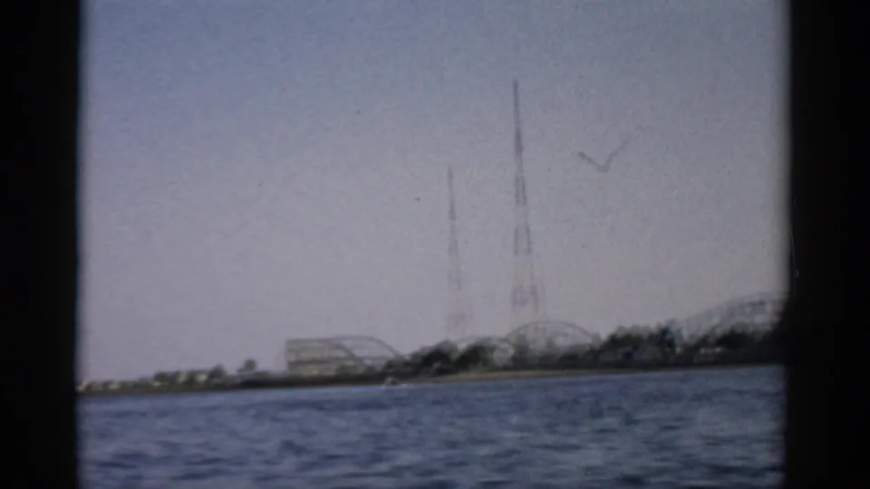8mm home-movie frame c.1957, Catskill-area lake, showing roller coaster and lattice towers on the far shore with a bird crossing the overcast sky.