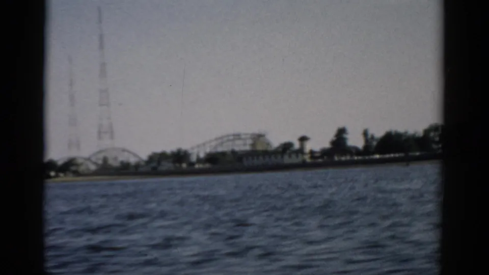 8mm frame from a 1957 Catskill-area boat ride, showing a tall lattice tower at left and roller coaster shoreline with apparent waterfront fencing.