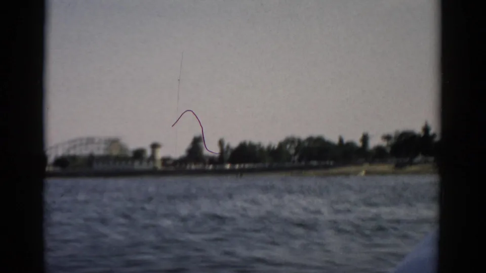 Late-clip 8mm frame c.1957, Catskill area, showing roller coaster and lattice tower on the far shore with a visible film-scratch artifact in the sky.
