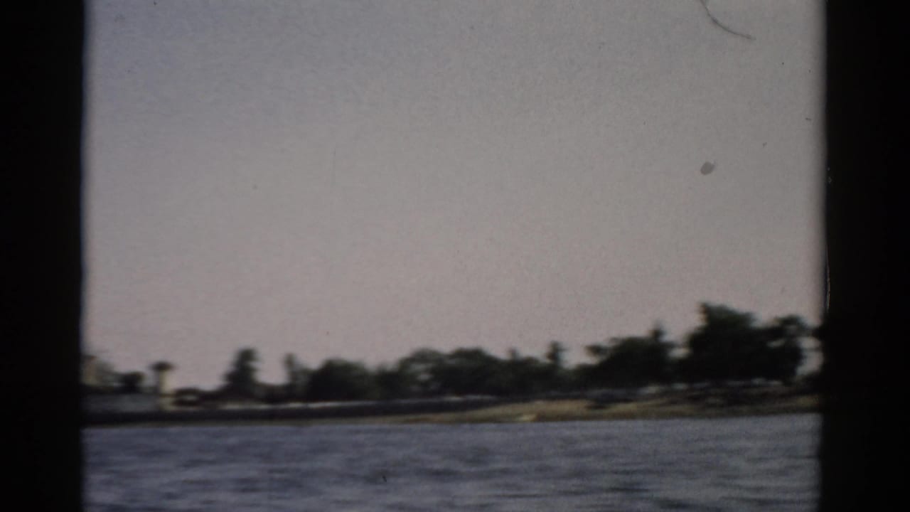 Older man in white shirt and dark cap smiling at camera from stern of a moving speedboat, boat wake visible behind him.